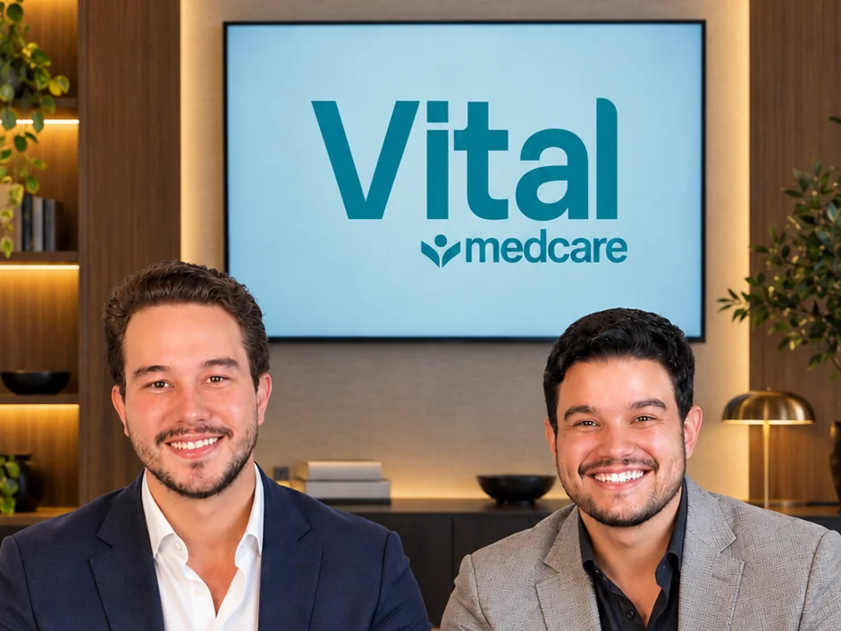 Os dois sócios fundadores da Vital sorrindo, sentados lado a lado e olhando para a câmera, com o logo da Vital numa TV ao fundo